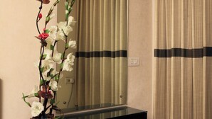 Un baño con espejo, un jarrón de madera con flores, un banco negro y cortinas color beige.