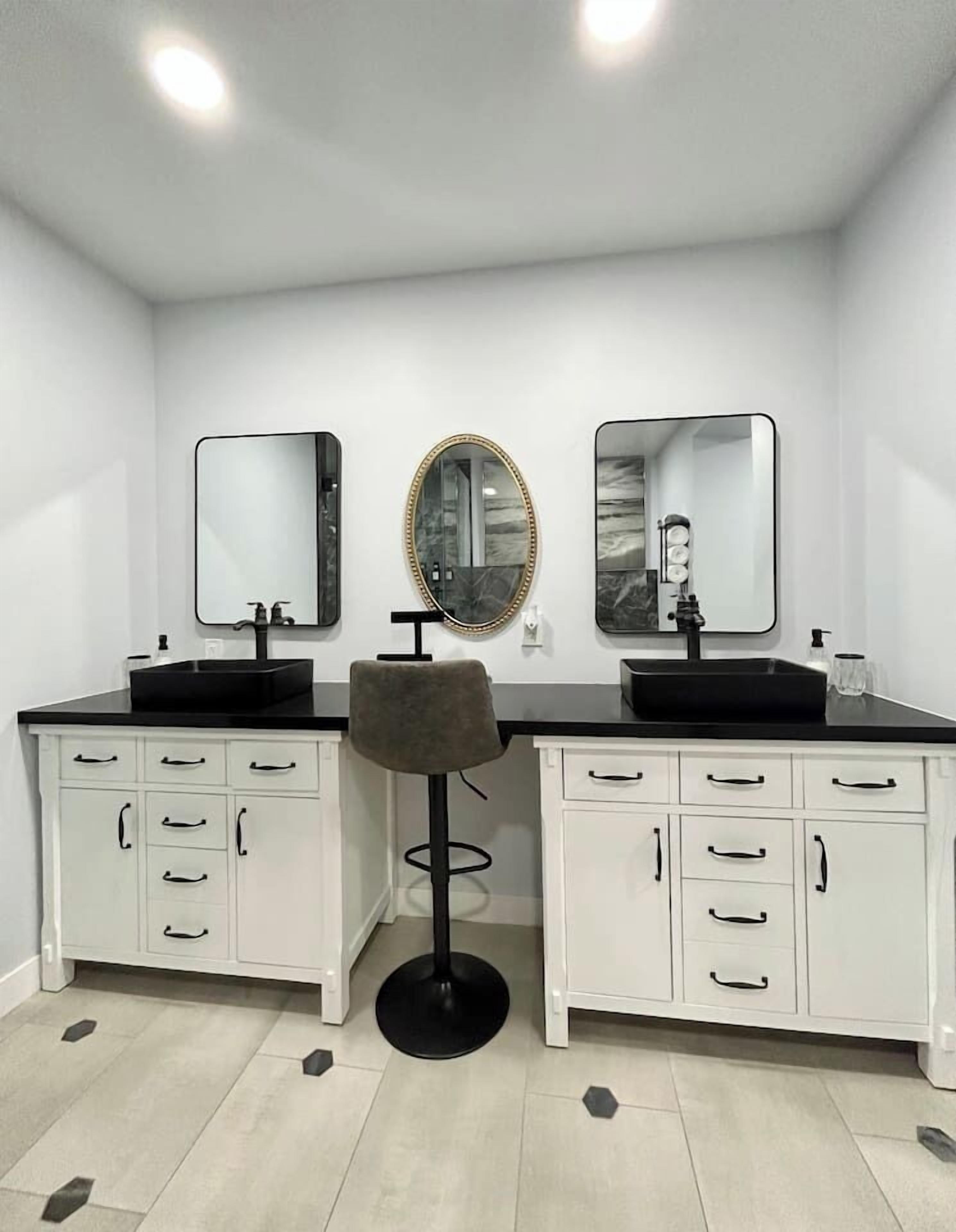 A bathroom with two sinks, two mirrors, a stool, and a white cabinet with drawers.