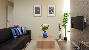 Una sala de estar con un sofá oscuro, una mesa de centro de madera, un jarrón con flores blancas, una planta en maceta y un televisor montado en la pared.