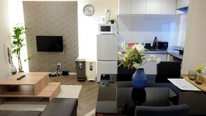 Una sala moderna con una televisión de pantalla plana, una mesa de centro, un sofá, una mesa de comedor con sillas, una zona de cocina con armarios y electrodomésticos, y un reloj en la pared.