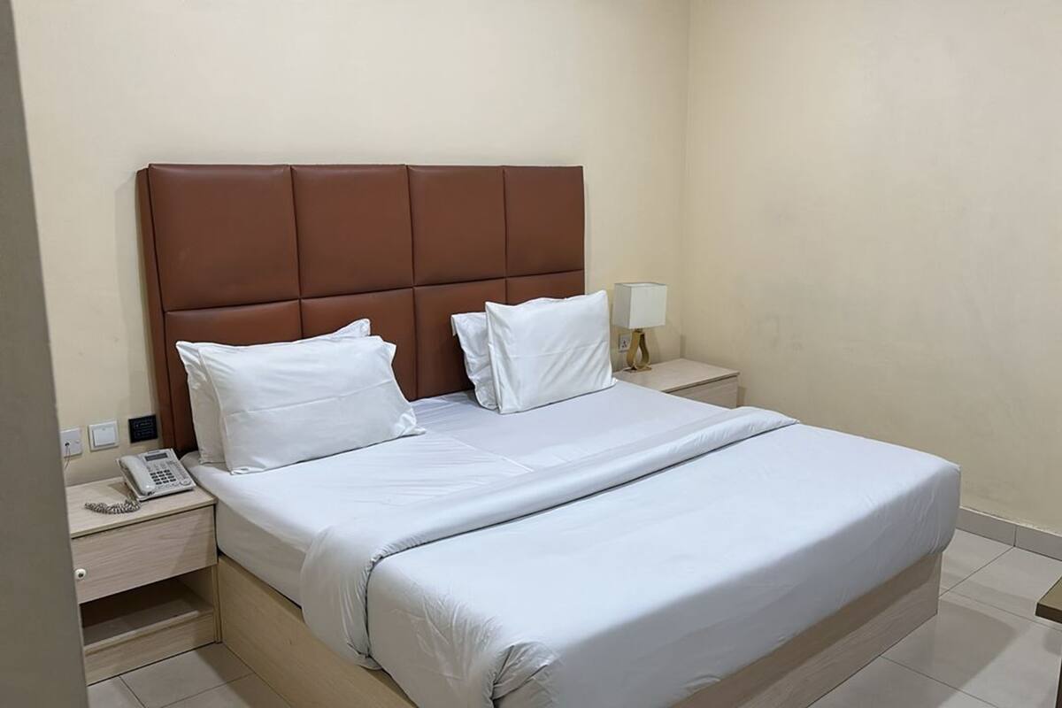 A hotel room with a bed, a nightstand with a phone, a lamp, and a chair.