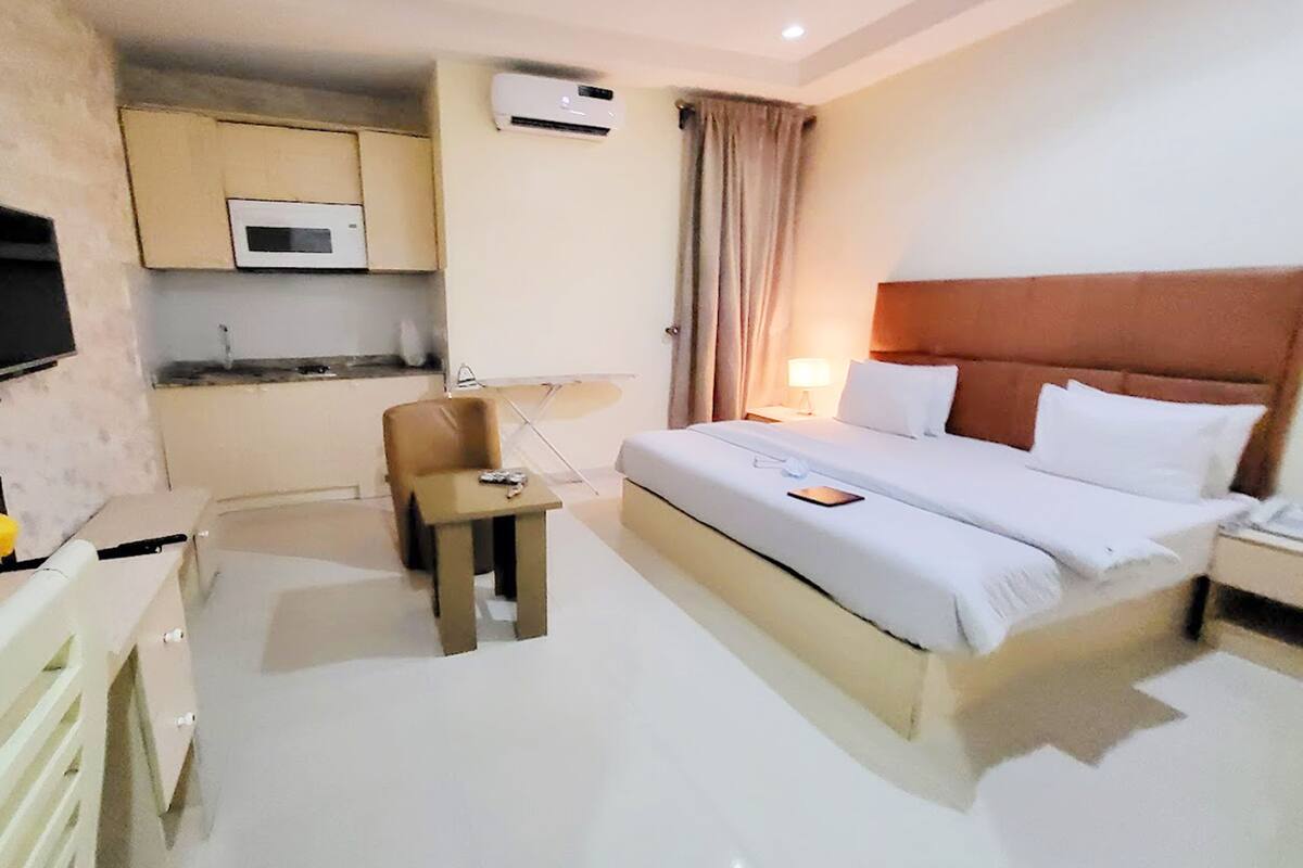 A hotel room with a bed, bedside table, lamp, small desk, chair, and a microwave in the kitchenette.