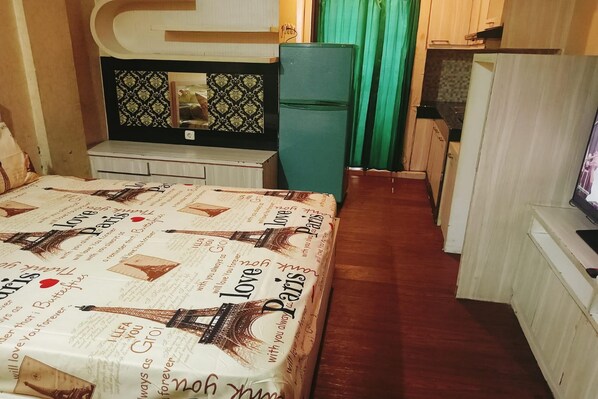 Quarto de hotel estreito com uma cama, uma pequena escrivaninha, um frigorífico e uma televisão.