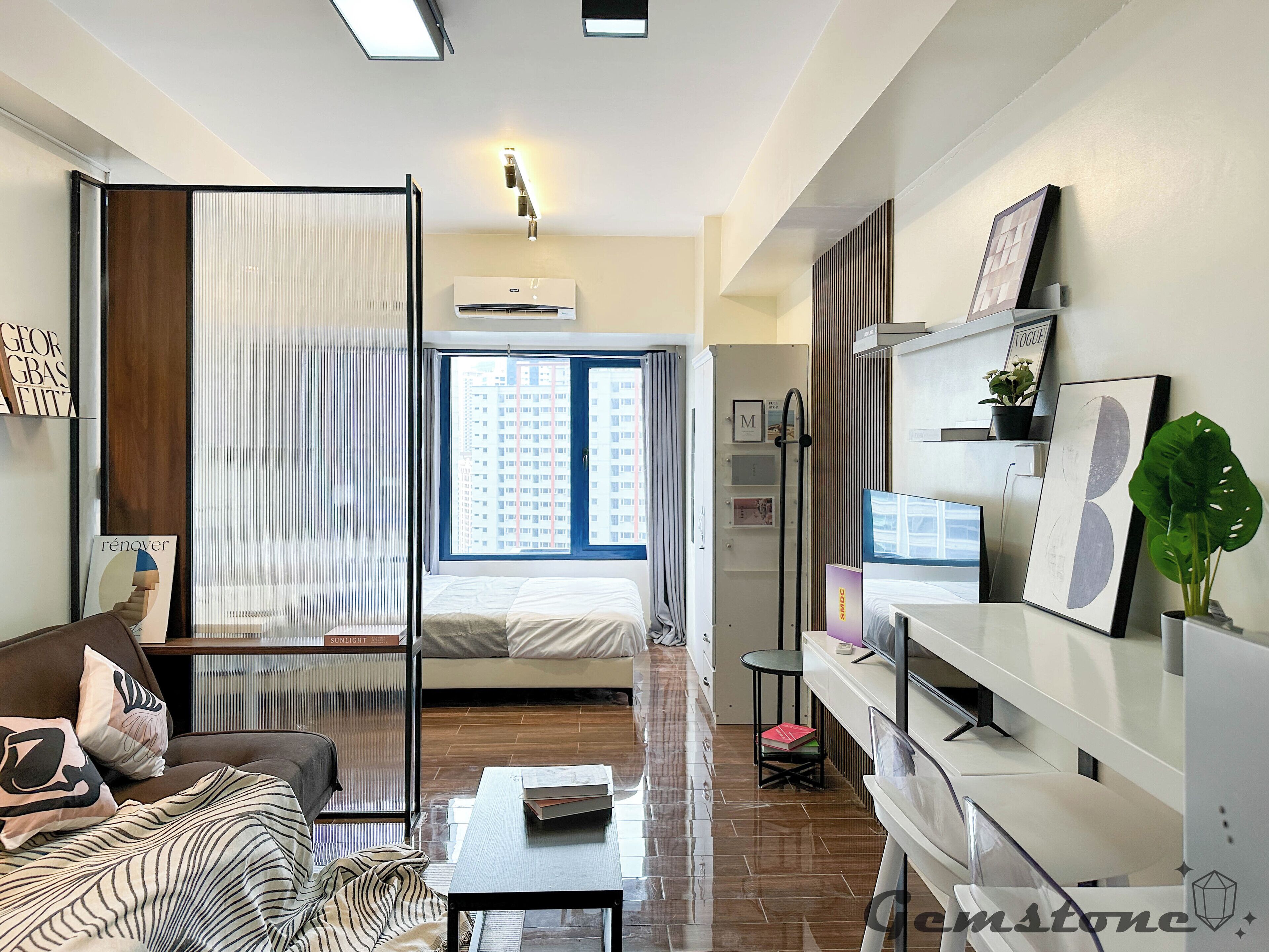 A modern hotel room with a bed, a sofa, a coffee table, a TV, and a large window with a city view.