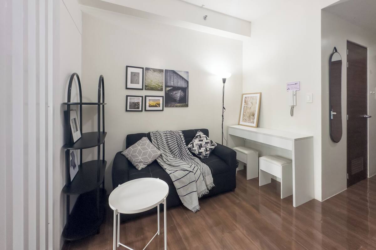 Una sala moderna con un sofá negro, una mesa de centro blanca y un escritorio blanco. Hay cuadros enmarcados en la pared, una lámpara de pie y una puerta a la derecha.
