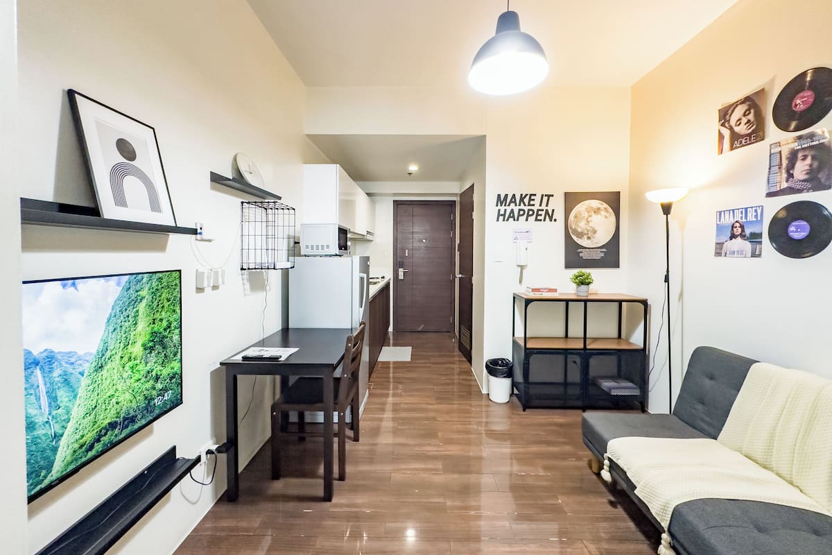 Una sala moderna con un televisor de pantalla plana, un sofá, una zona de comedor con mesa y sillas, una cocina con microondas, y un pasillo que conduce a una puerta.