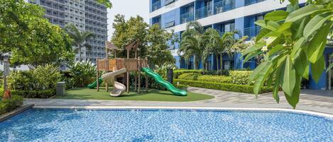 Una piscina con zona de juegos infantiles, rodeada de vegetación y edificios modernos.