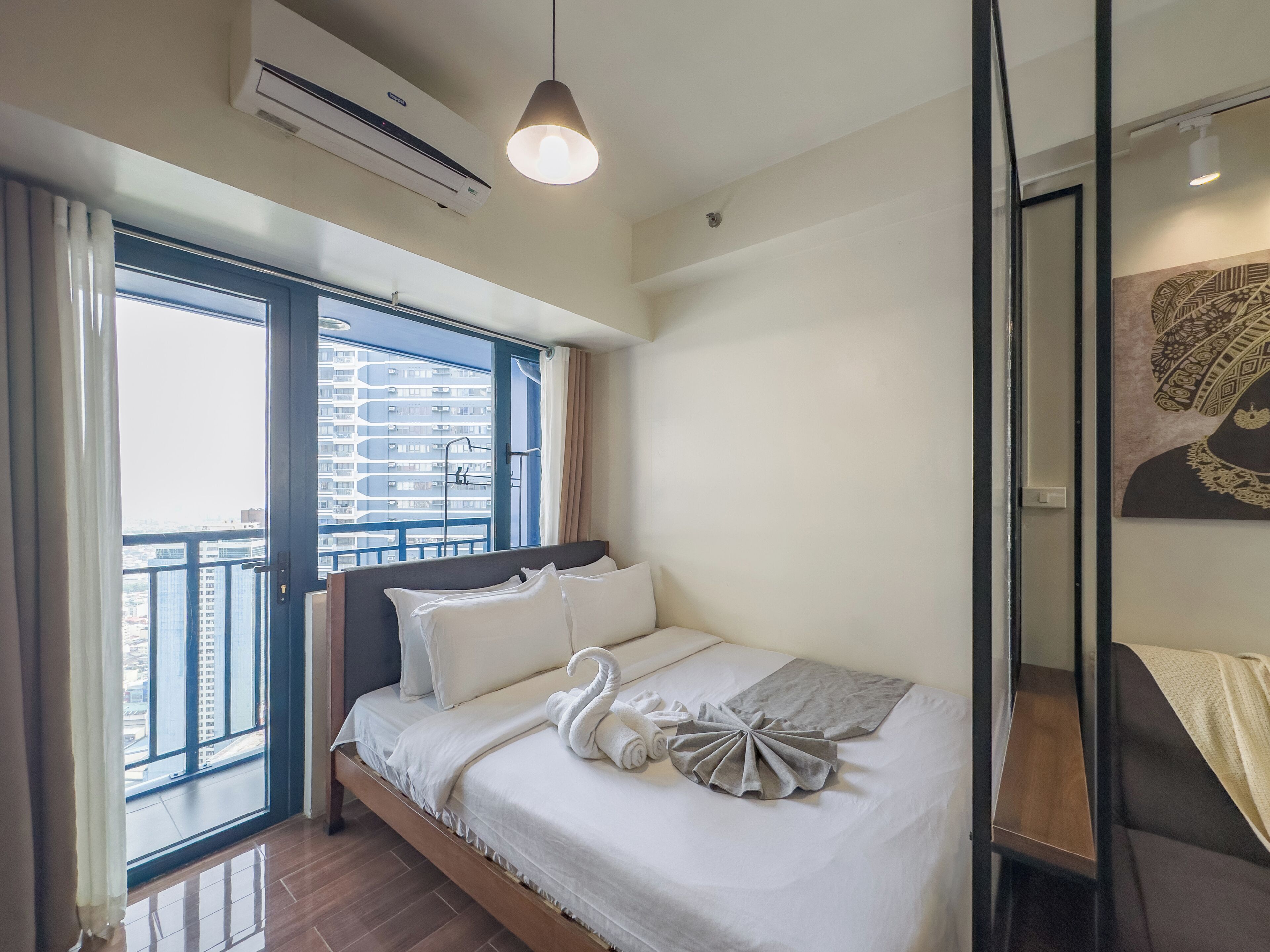 Une chambre d’hôtel moderne dotée d’un grand lit, offrant une vue sur la ville grâce à de larges fenêtres du sol au plafond, et d’un climatiseur fixé au plafond.