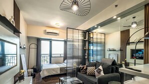 A modern hotel room with a bed, a sofa, a small table, and a mirror. There is a window with a view of the city. - GemstoneBR in SMDC Air Residences (Makati)