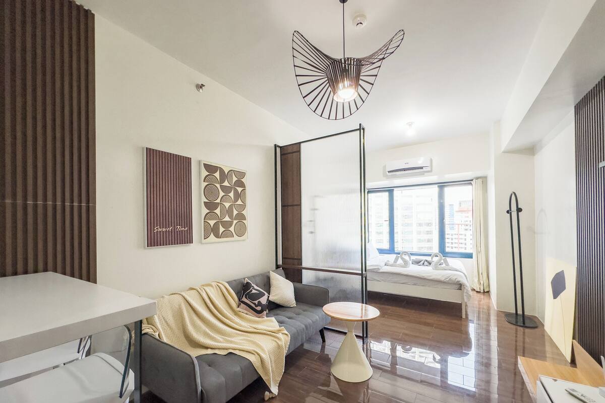 Una habitación de hotel moderna con un sofá gris, una pequeña mesa redonda, un espejo grande y una lámpara de techo original.