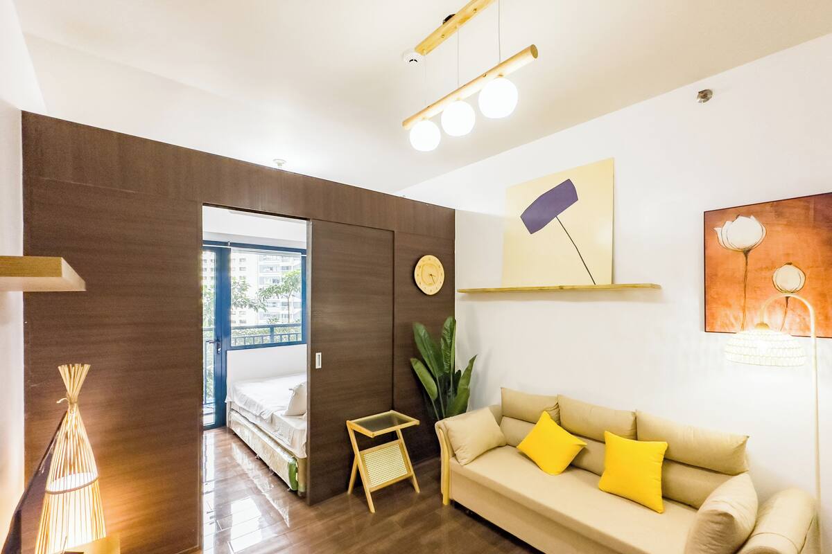 Una sala moderna con un sofá, una mesita y una planta. Una puerta da a un balcón con vistas a edificios.