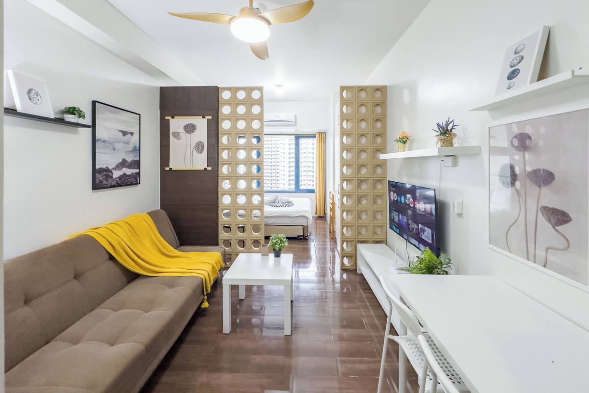 Una sala de estar moderna con un sofá marrón, una mesa de centro blanca, un ventilador de techo y un televisor sobre un mueble blanco.