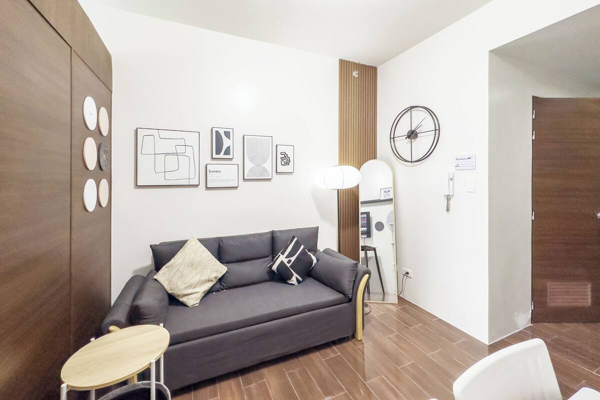 Una sala de estar moderna con un sofá gris oscuro, una mesita redonda y un espejo. Se observan obras de arte enmarcadas en la pared y un panel de madera a la izquierda.