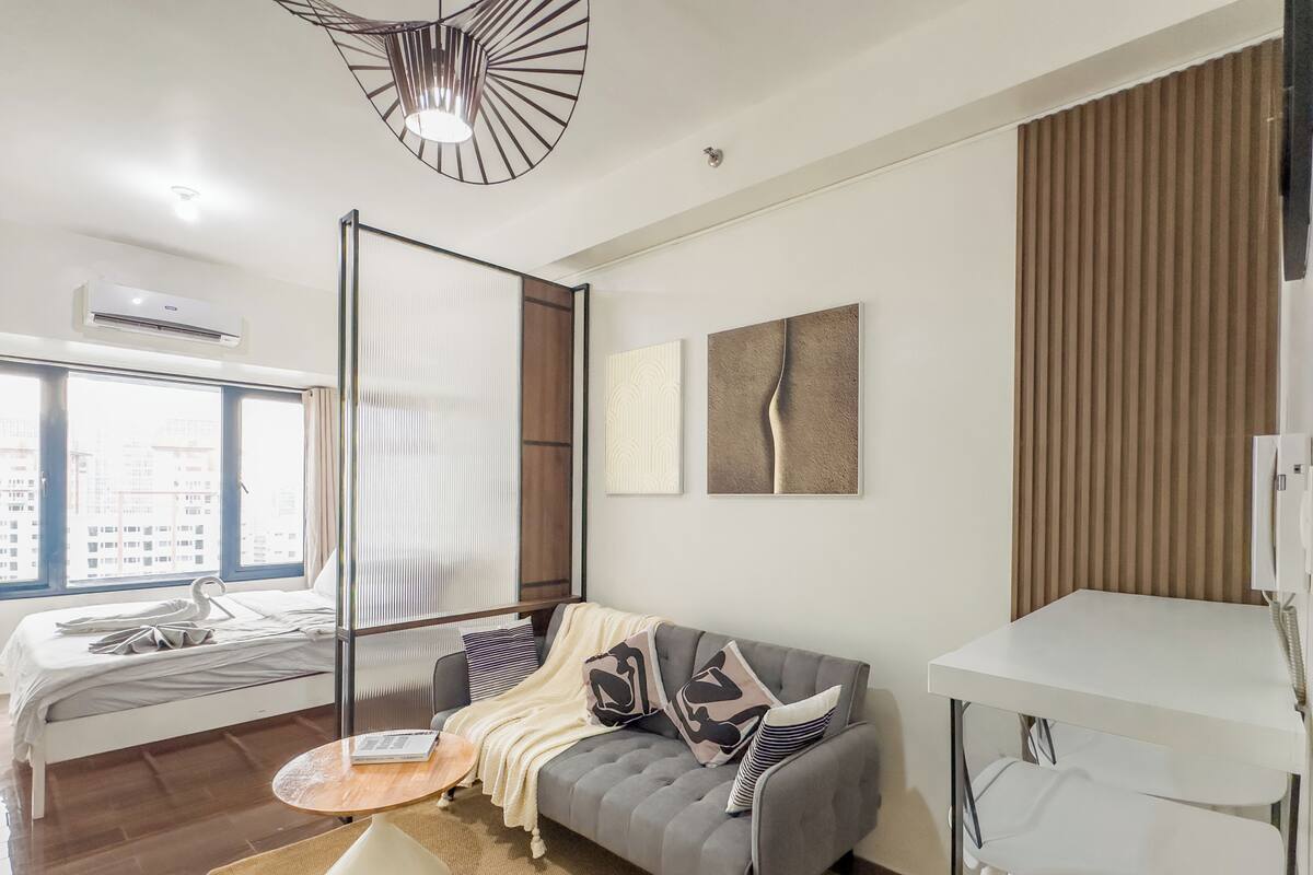 Una habitación de hotel moderna con una cama, un sofá gris, una mesita redonda y un escritorio blanco.