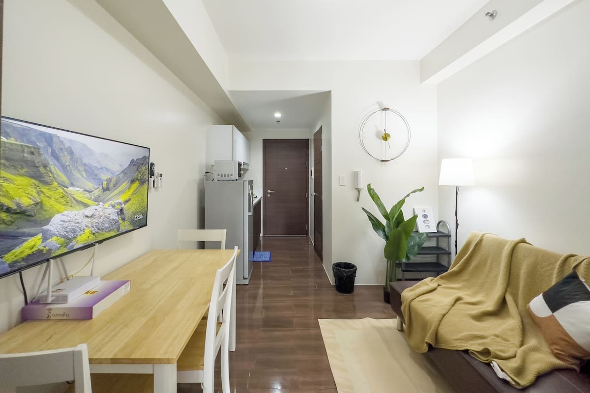 Un moderno salón con una mesa de comedor de madera, sillas blancas, un sofá con una manta, un televisor de pantalla plana grande, una planta y un reloj de pared.