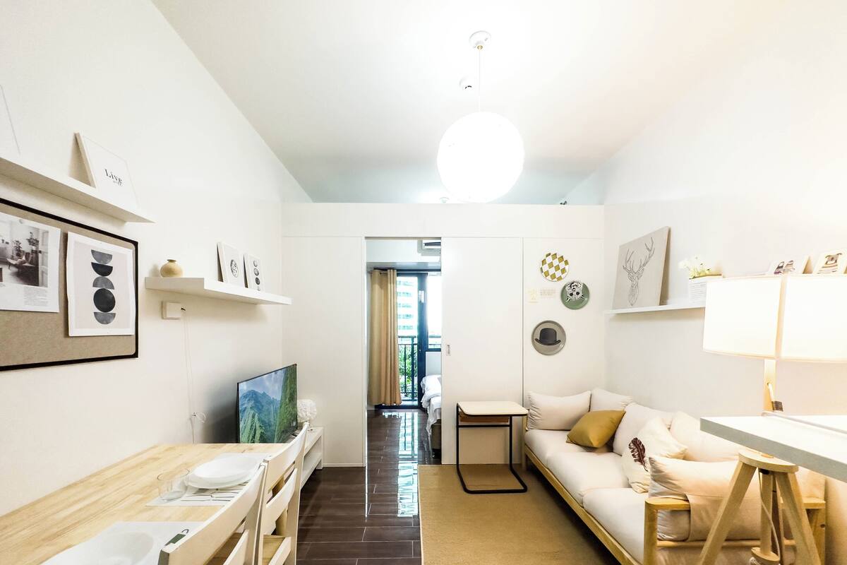 Una sala de estar moderna y bien iluminada, con un sofá, una mesa de comedor y un televisor. Hay cuadros enmarcados en la pared y una mesita con una lámpara.