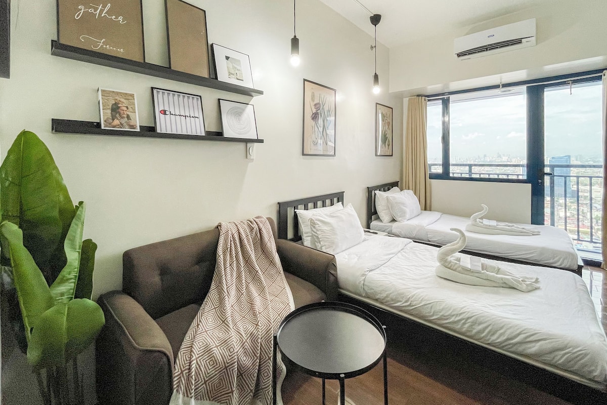 Habitación de hotel con dos camas individuales, un sofá, una mesita redonda y un ventanal con vistas a la ciudad.