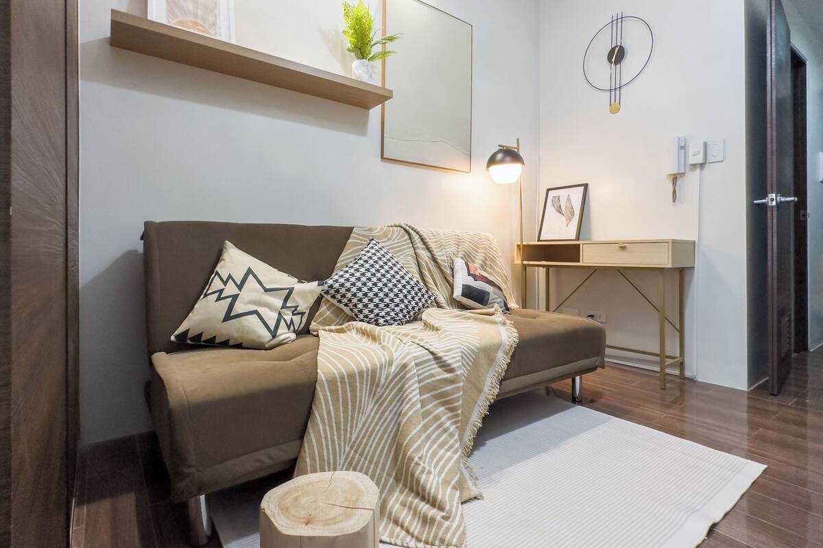 Una sala de estar moderna con un sofá marrón, cojines estampados, un taburete de madera, una alfombra blanca y una decoración minimalista que incluye una planta, una obra de arte enmarcada y un reloj de pared.