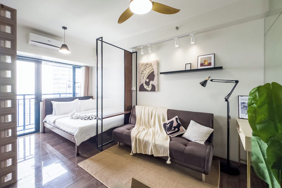 Habitación de hotel moderna con cama, sofá, ventana grande, ventilador de techo y planta.