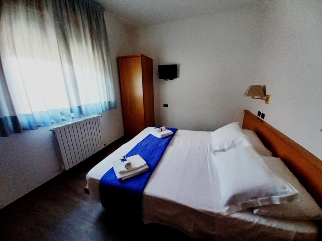 Hotellihuone, jossa on sänky, sininen pyyhe, puinen vaatekaappi, seinälle asennettu televisio ja ikkuna, jossa on verhot.