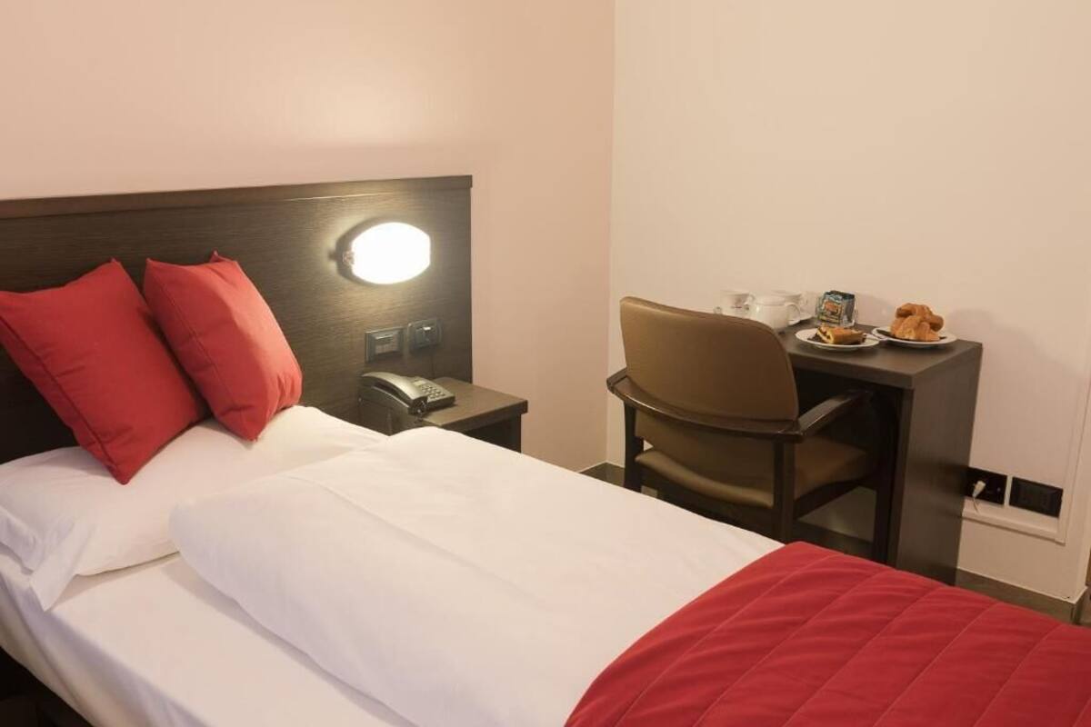 A hotel room with a bed, a desk with a chair, a phone, and a small table with breakfast items.