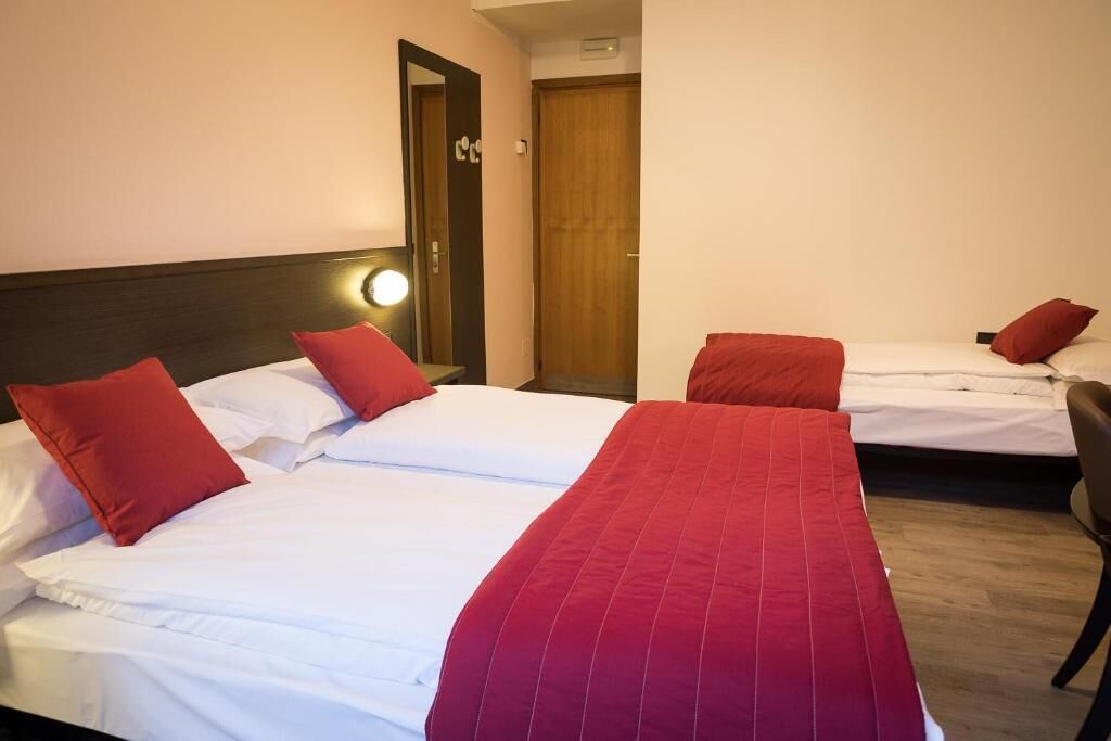Habitación de hotel con dos camas, cada una con sábanas blancas y almohadas rojas. Se observa una puerta de madera y una pequeña mesa con una lámpara.