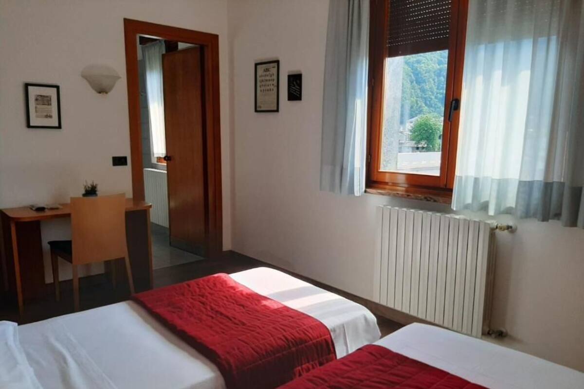 A hotel room with two beds, a desk, a chair, a radiator, and a window with curtains.
