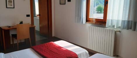 A hotel room with two beds, a desk, a chair, a radiator, and a window with curtains.