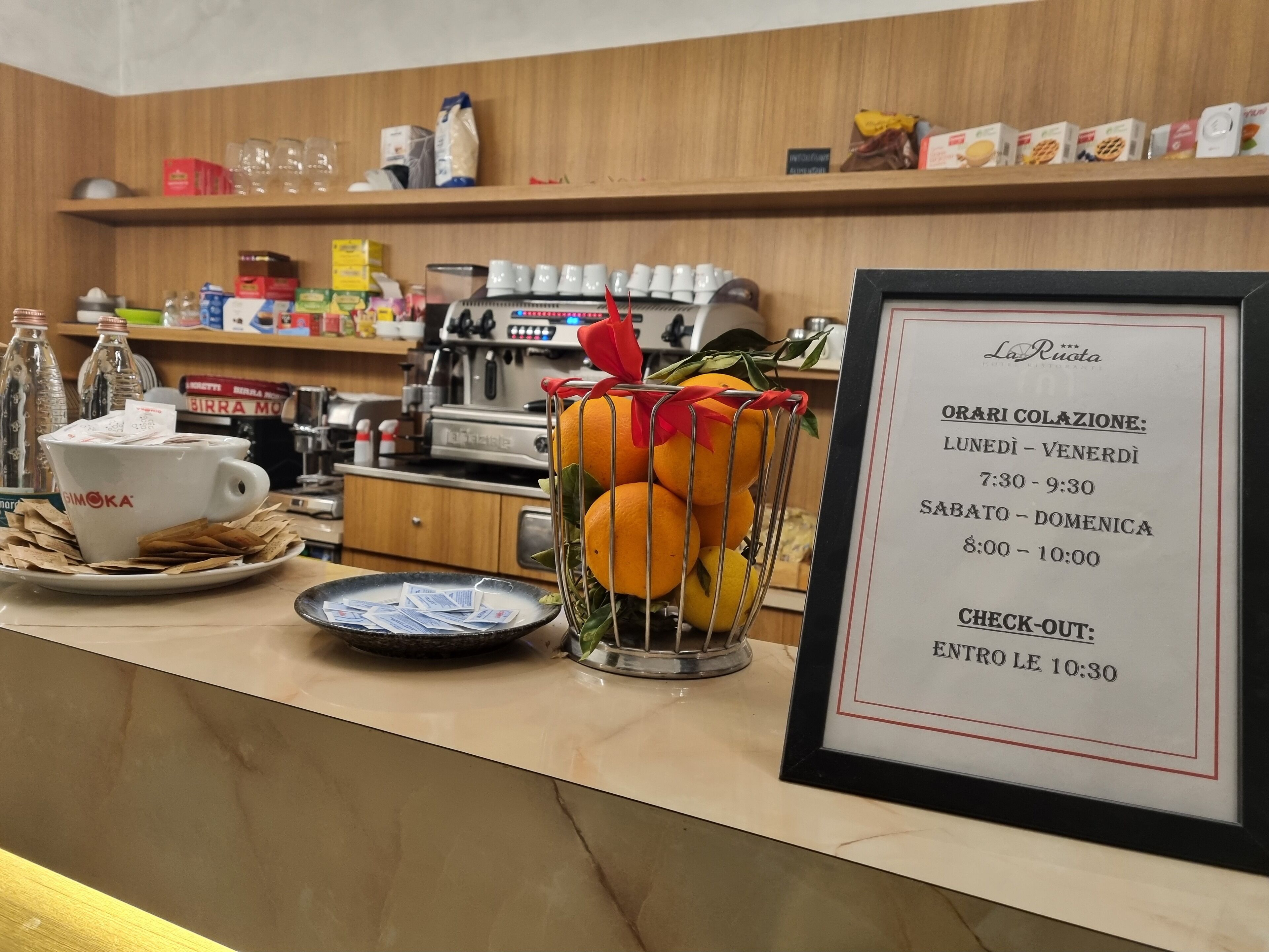 Un espace d’accueil d’hôtel avec un coin café, un tableau affichant le menu et divers appareils de cuisine en arrière-plan.