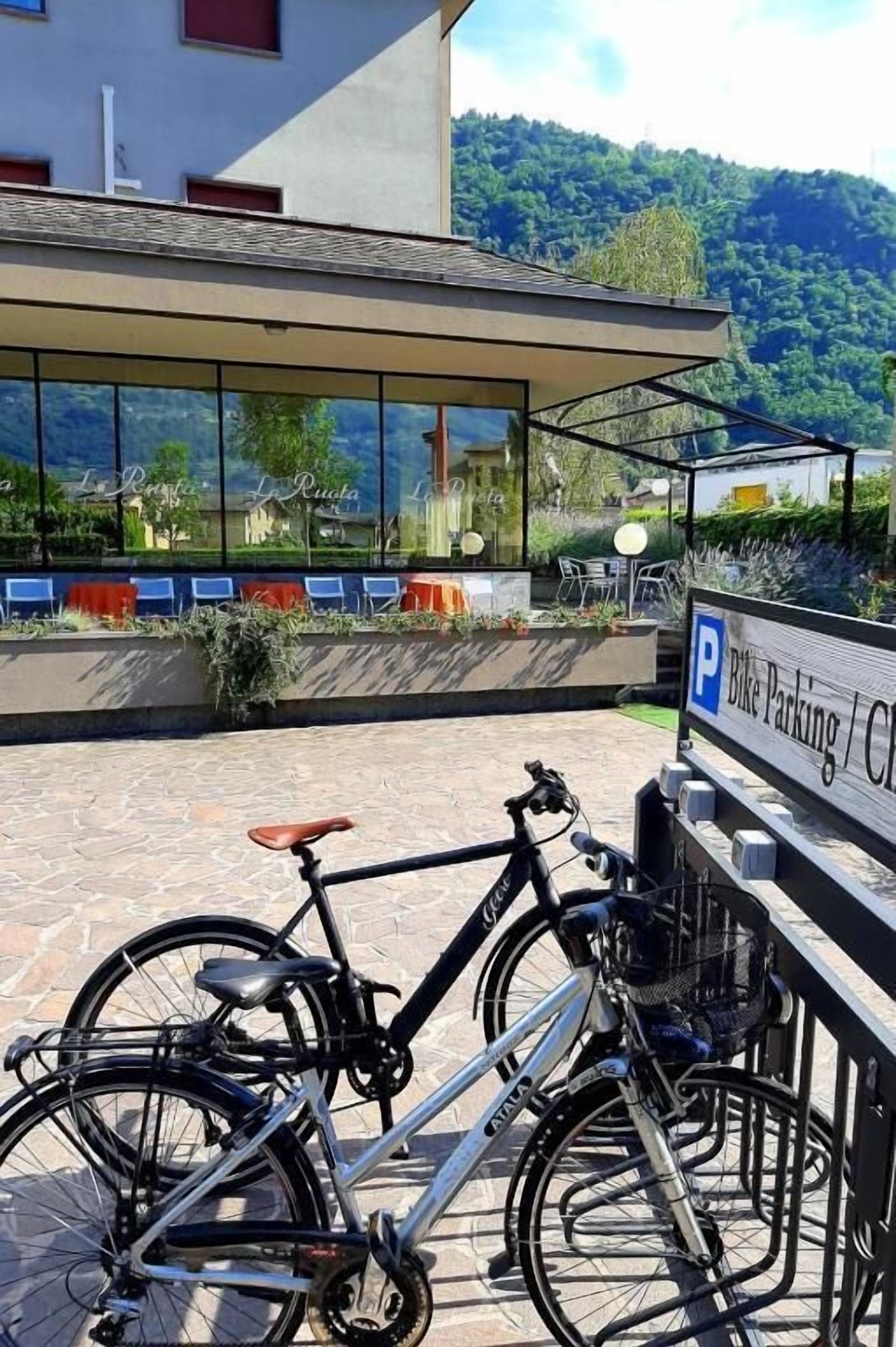 Bicicleta estacionada em suporte, em frente a um edifício com vista para uma montanha e área de estar ao ar livre com mesas.