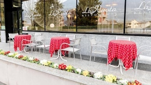 An outdoor dining area with tables covered in red checkered tablecloths, chairs, and a flower bed in front. - Hotel La Ruota (Morbegno)