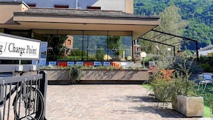 A modern building with a stone-paved entrance, a covered outdoor seating area, and a sign indicating a charge point for electric vehicles. - Hotel La Ruota (Morbegno)