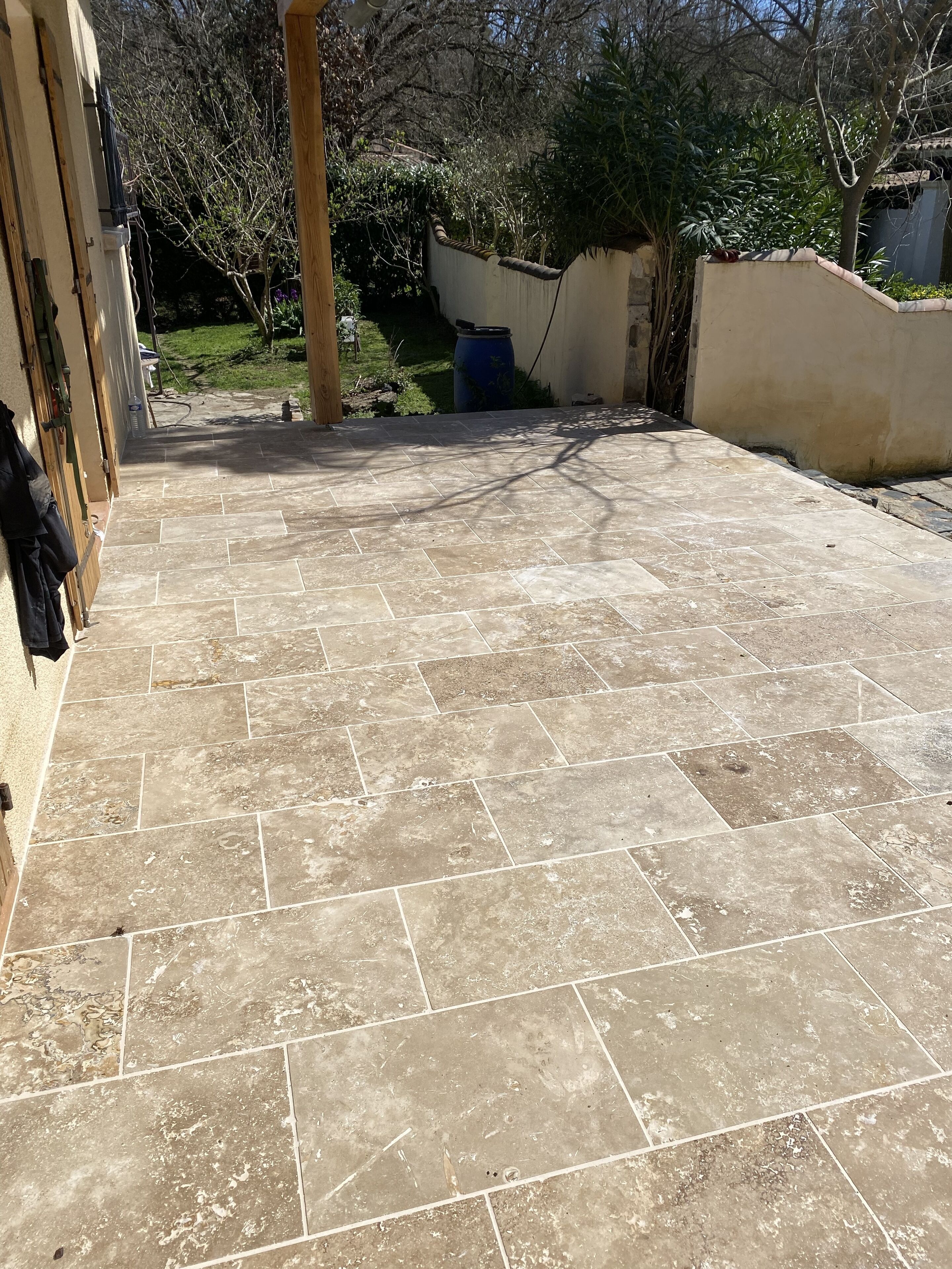 A patio with tiled flooring, a wooden structure on the left, and a garden in the background.