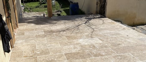 A patio with tiled flooring, a wooden structure on the left, and a garden in the background.