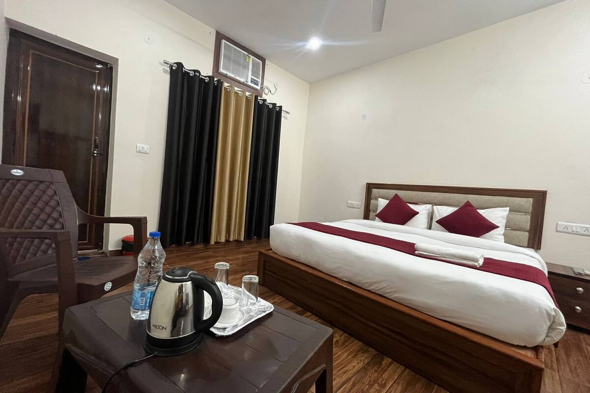 A hotel room with a bed, a table with a kettle and water bottle, a chair, and curtains.