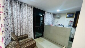 A living room with a sofa, a glass door leading to a balcony, and a kitchenette area with a fan and calendar. - Badal Inn (New Delhi)