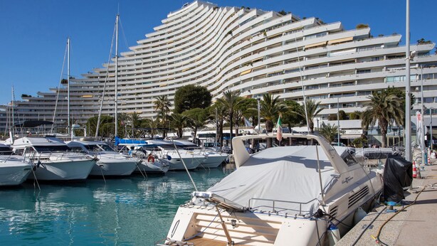 Een jachthaven met diverse jachten afgemeerd, een groot modern gebouw met balkons en palmbomen op de achtergrond.