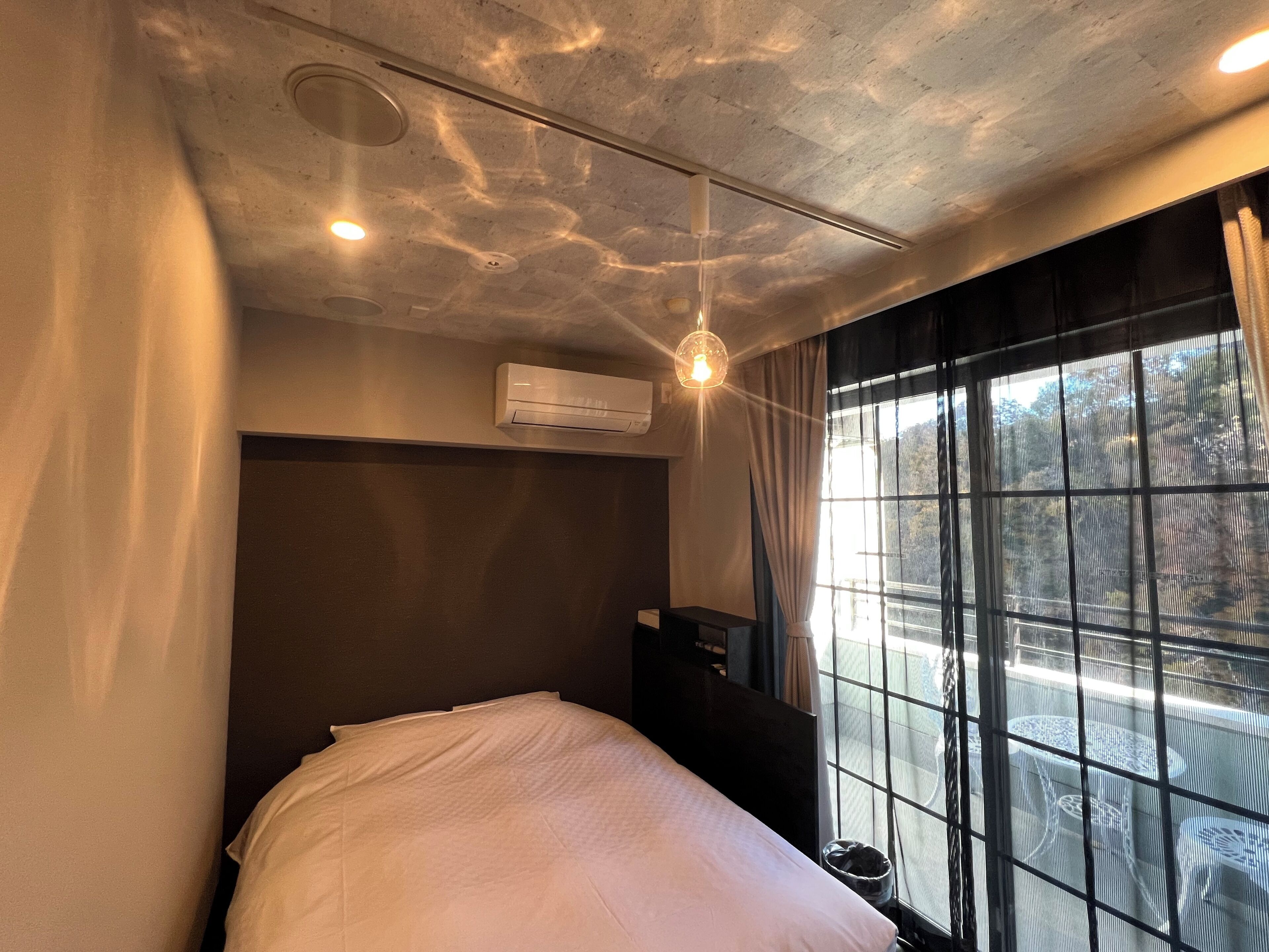 A hotel room with a bed, an air conditioning unit, a desk, and a window with a view of trees.