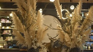 Une pièce avec des poutres apparentes en bois au plafond, un luminaire central et une composition décorative composée d’herbe des pampas et d’une sculpture de cerf, le tout disposé sur une table en bois.