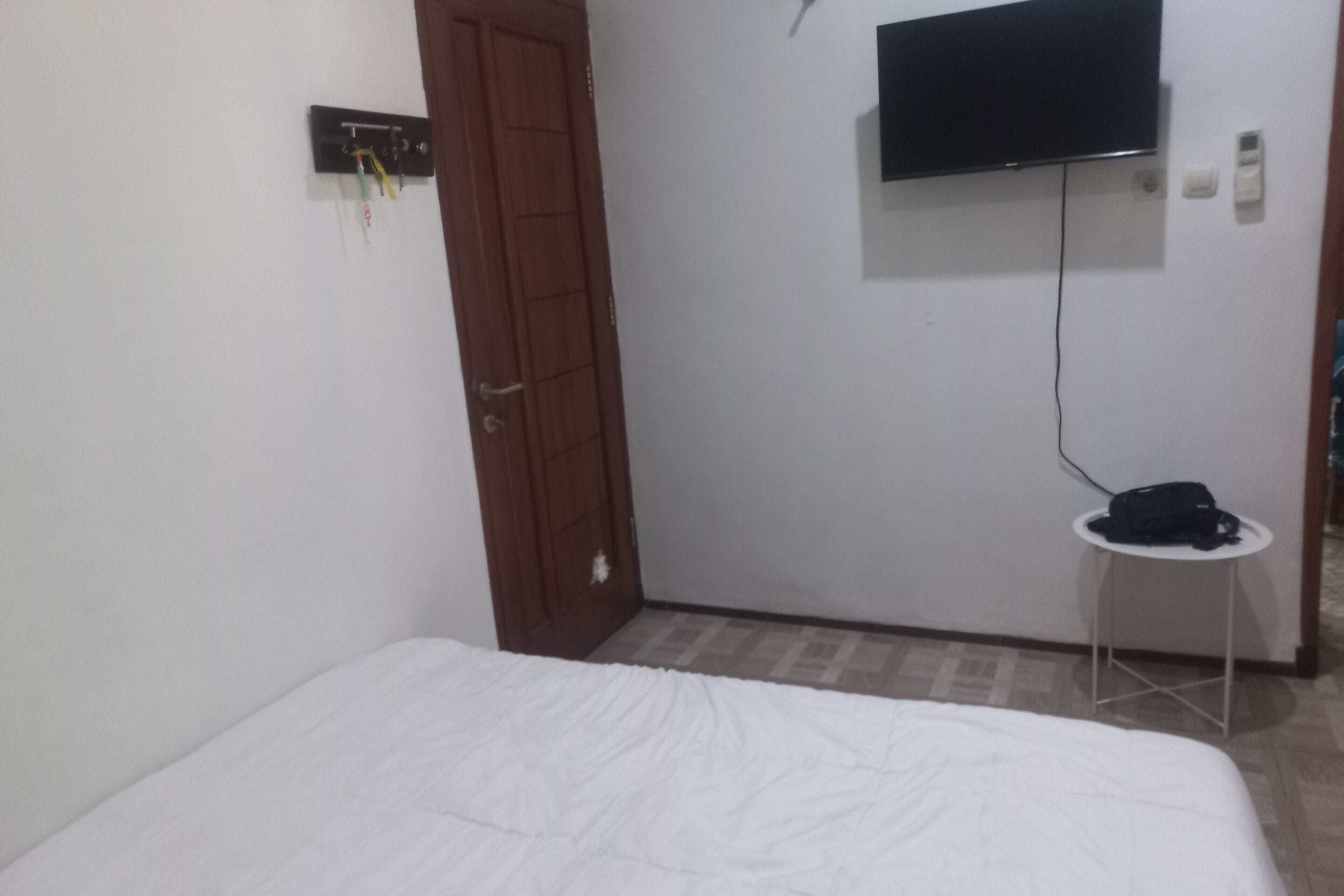 A room with a bed, a television mounted on the wall, a small table with a bag on it, and a door.