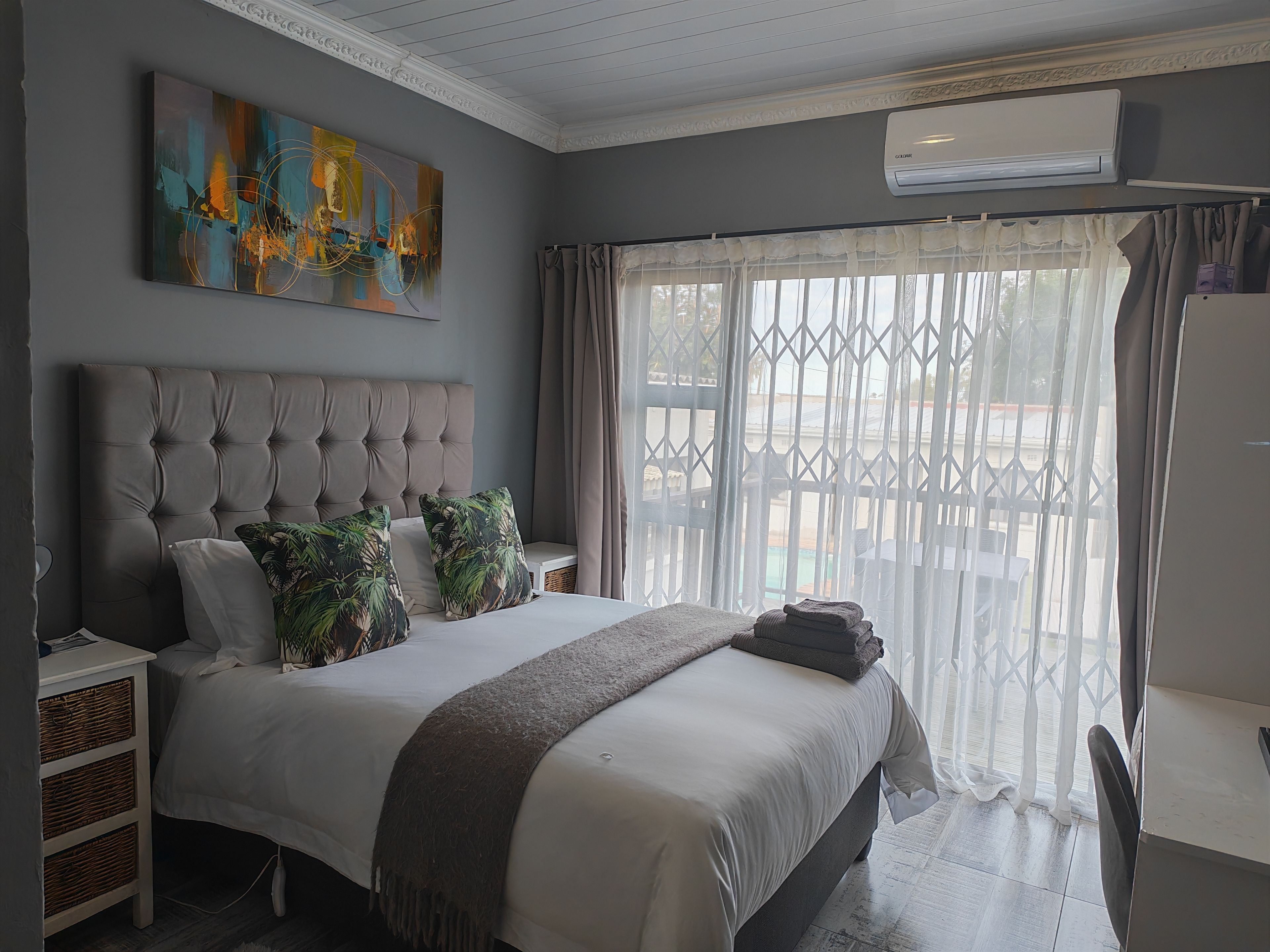 Quarto com cabeceira estofada, uma grande pintura abstrata, cama com edredom branco e vista pela janela com cortinas leves.