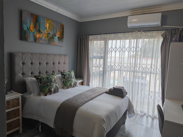 A bedroom with a tufted headboard, a large abstract painting, a bed with a white comforter, and a view through a window with sheer curtains.