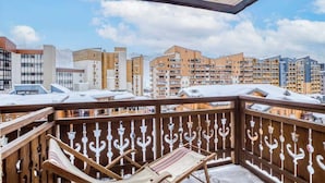 Un balcon doté de balustrades en bois et de deux fauteuils de jardin, offrant une vue sur un paysage urbain avec de nombreux immeubles et un ciel dégagé.