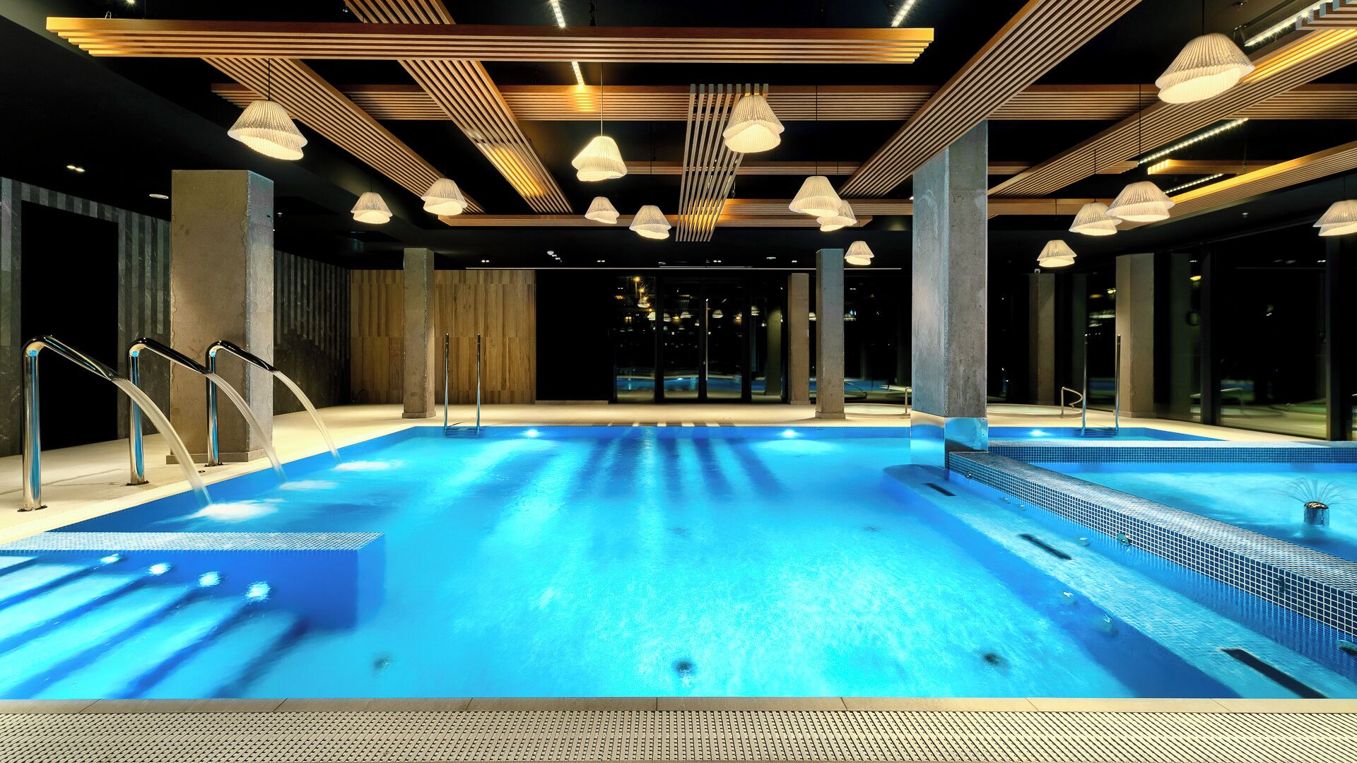 A large indoor swimming pool with clear blue water, surrounded by wooden beams and modern lighting fixtures.