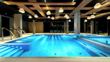 A large indoor swimming pool with clear blue water, surrounded by wooden beams and modern lighting fixtures.