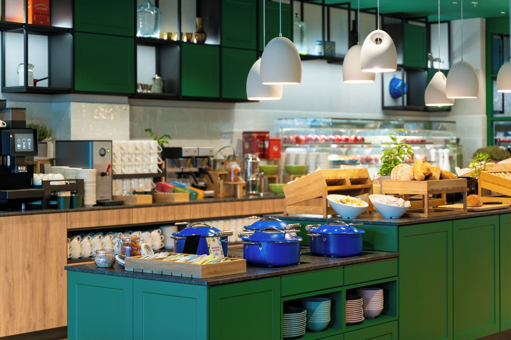 Una cocina moderna con armarios verdes, una mesa de buffet con ollas azules, y lámparas colgantes.