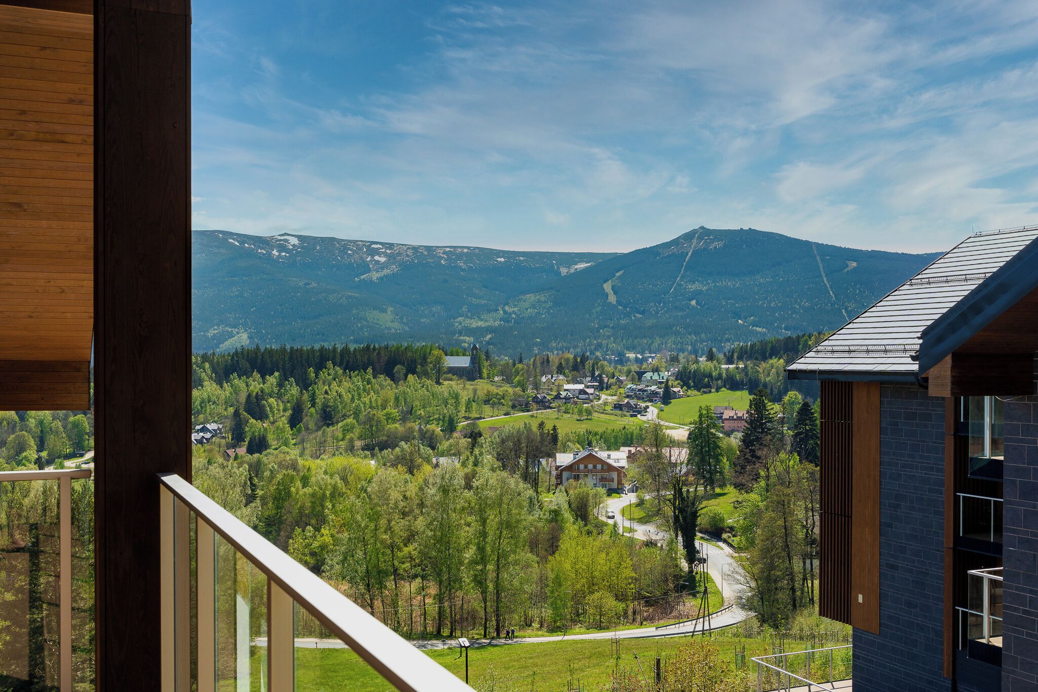 Vista de la montaña desde un balcón con barandilla de madera, con vistas a un valle con casas y vegetación.