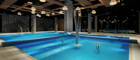 A modern indoor swimming pool with a blue tiled floor, multiple light fixtures, and a dark ceiling with wooden beams.