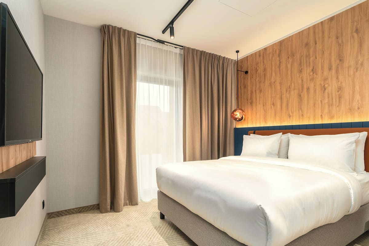 Ett hotellrum med en stor säng, en platt-TV, en sänggavel i trä och ett stort fönster med gardiner.