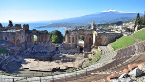An ancient amphitheater with stone seating and ruins, overlooking a coastal town and a mountain.